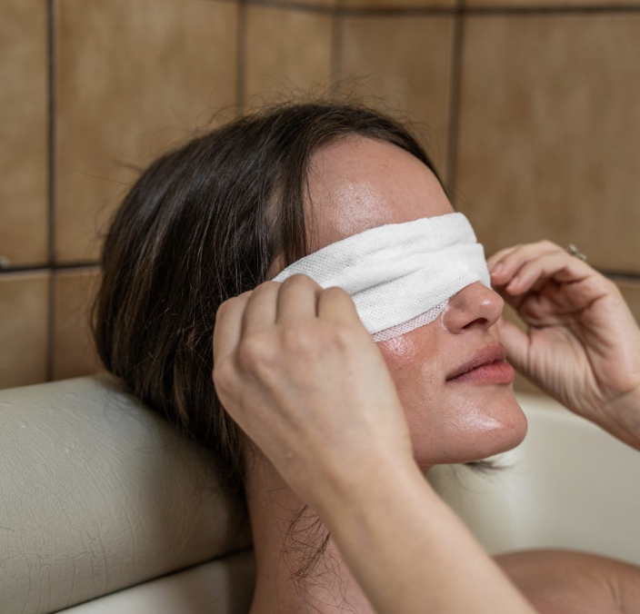 A woman sits in a bathtub and covers her eyes with a white towel, gently holding each side. The background features beige tiled walls. Fearrington Village